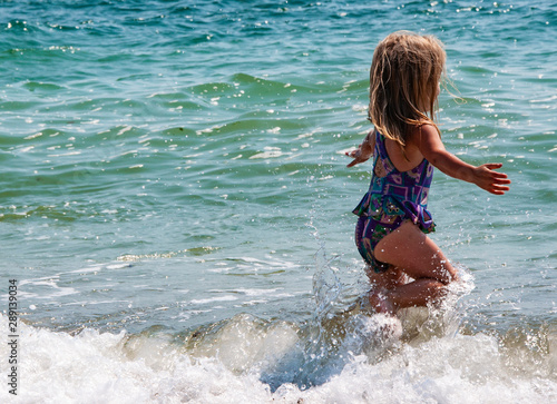 Cute happy little girl running along the beach in swimming suit, jumping over waves. Beautiful summer sunny day, blue sea