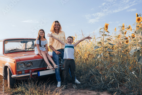 Happy family near sunflowers field, mom with two children, girl teen in dress sitting on hood of the red retro car, mother and son standing beside, summer vacation © Vasya