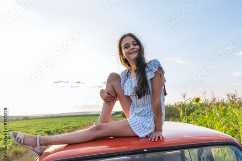 Happy cute teen girl in striped blue dress sitting on the roof of the red retro car among the field, smiling and looking at the camera, blue sky on the background