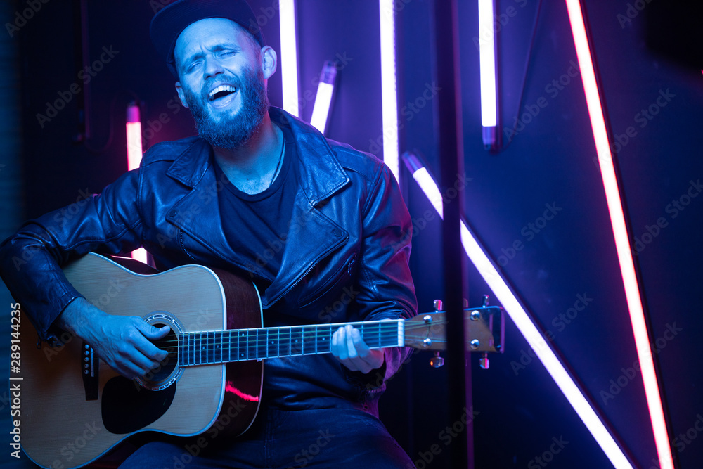 Obraz premium Singer and guitar player singing on a stage with neon lights. He is a rocker and he is wearing leather biker jacket or asymmetric zip jacket with black cap, jeans