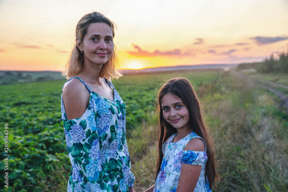 Close up portrait of beautiful young mom with her teen brunette daugther, both in dresses with flowers print standing among the mealow and looking at the camera, sunset on the background