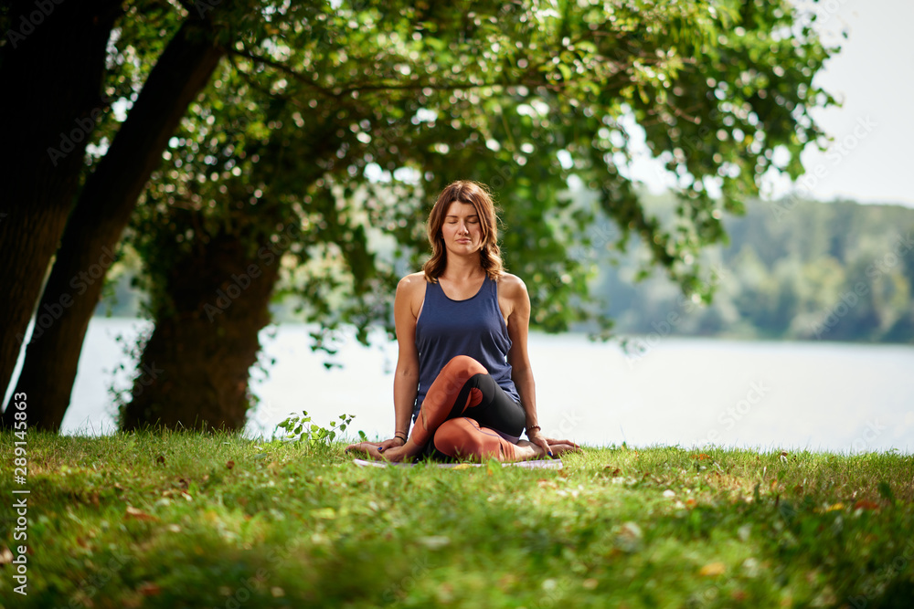 Beautiful Cucasian brunette dressed in sports clothing sitting on mat in nature in knee pile yoga pose.