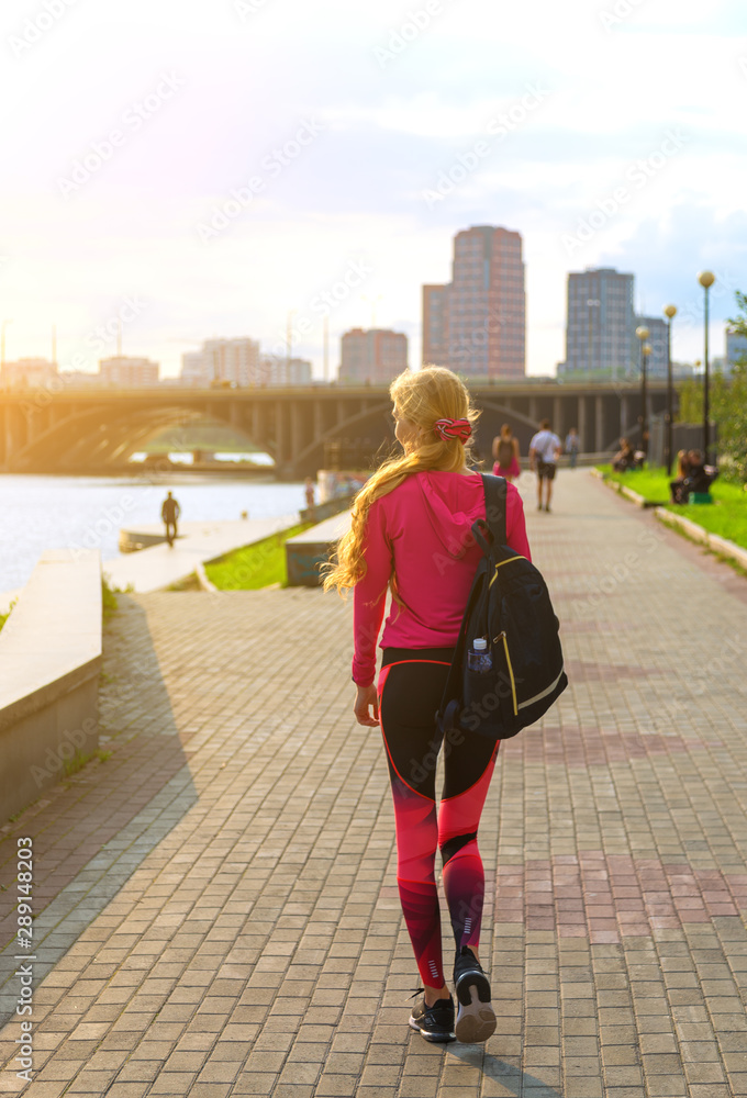 sporty girl with backpack walks on the waterfront at sunset