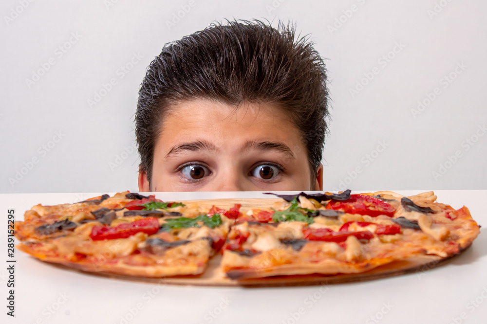 The boy eats Italian pizza. Looks out from behind the table The boy eats Italian pizza. Looks out from behind the table