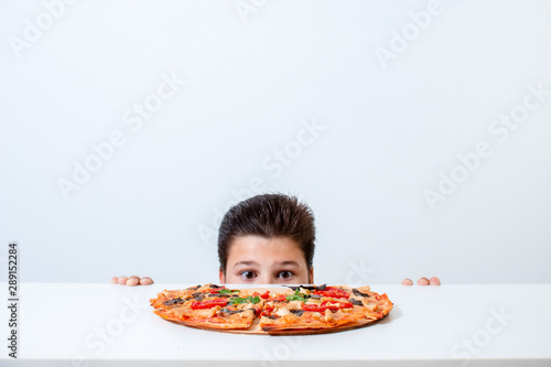 The boy eats Italian pizza. Looks out from behind the table