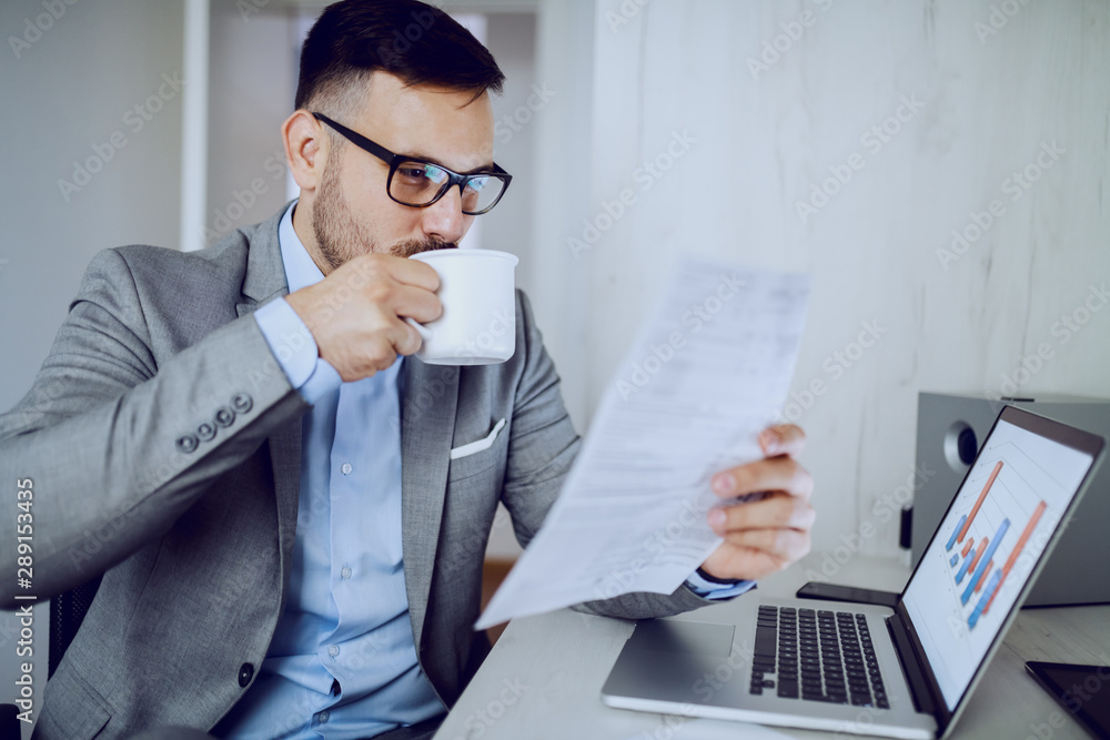 Focused hardworking manager in gray suit and with eyeglasses reading report and drinking coffee while sitting in office. On desk is laptop. Focused hardworking manager in gray suit and with eyeglasses reading report and drinking coffee while sitting in office. On desk is laptop.