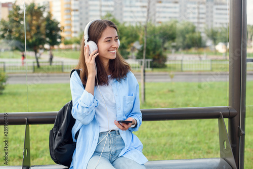 Teen girl listens to the music by white headphones in a public transport station while she waiting for tram. © gorynvd
