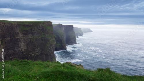 Photograph of the Cliff of Moher, overlooking the meadow and the sea. In the background clouds and mist.