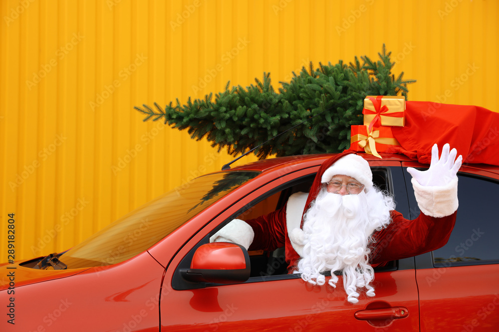 Obraz premium Authentic Santa Claus with fir tree and bag full of presents driving car against yellow background