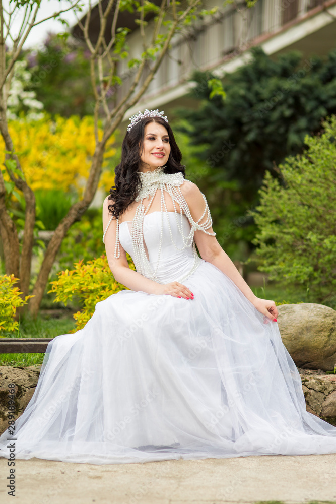 Portrait of Sensual Caucasian Bride With Diadem Sitting In Flowers Garden Outdoors. Wearing Beads Necklace.