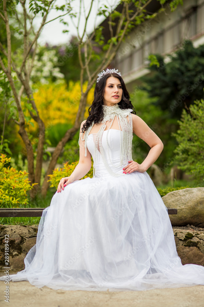 Portrait of Caucasian Bride With Diadem Sitting In Flowers Garden Outdoors. Wearing Beads Necklace. Portrait of Caucasian Bride With Diadem Sitting In Flowers Garden Outdoors. Wearing Beads Necklace.