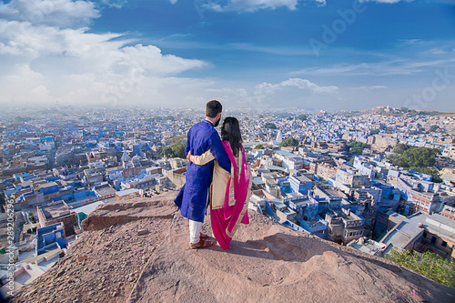 Asian indian couple standing together on the hill wearing traditional ethnic dress looking at out with blue house in the background, blue city jodhpur - travel concept