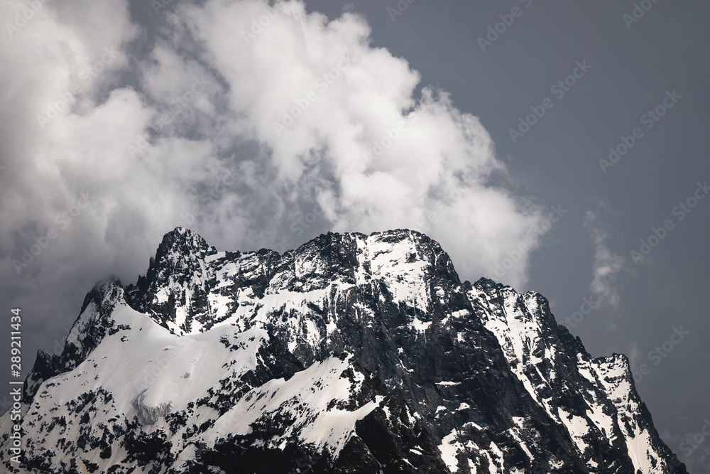 View of a high steep rocky mountain partially covered with snow against a dark cloudy sky with clouds clinging to the top. Mountain weather change concept
