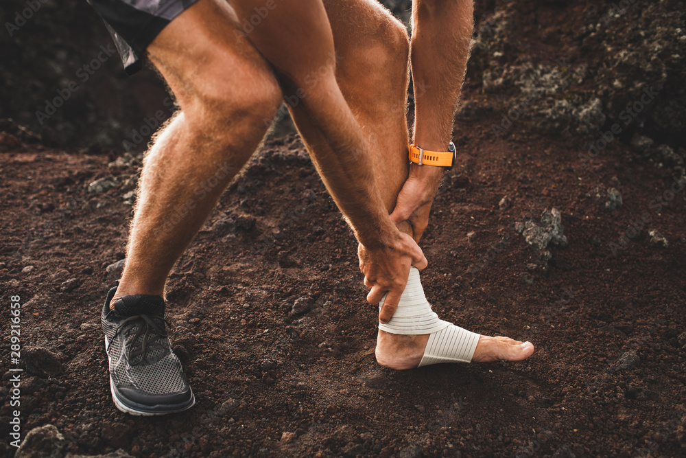 Male runner holding injured leg close-up and suffering with pain. Leg injury. Sprain ligament or tendon while running outdoors. Compression bandage on ankle. Male runner holding injured leg close-up and suffering with pain. Leg injury. Sprain ligament or tendon while running outdoors. Compression bandage on ankle.