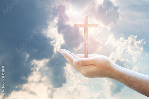 Fotografia, Obraz Human hands holding wooden cross against the light from cloudy blue sky, Christi