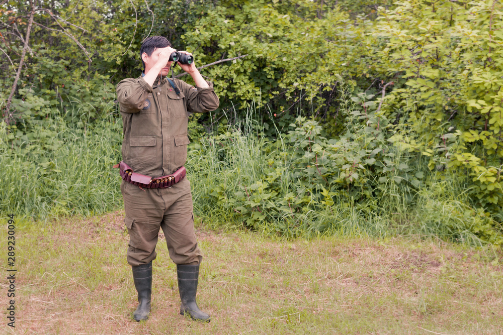 A male hunter with binoculars looks out for his prey against the background of the forest. The concept of a hunter, successful or failed hunt. Image.