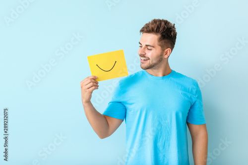 Happy young man with drawn smile on sheet of paper against color background