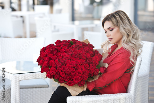 Elegant beautiful blonde woman is wearing green fashion dress in cafe holding with big bouquet of 101 red roses. Birthday.