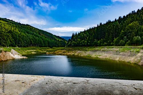 Image of an old concrete fence and lake with the beauty of the blue sky and the slopes covered by the green forest in the background.