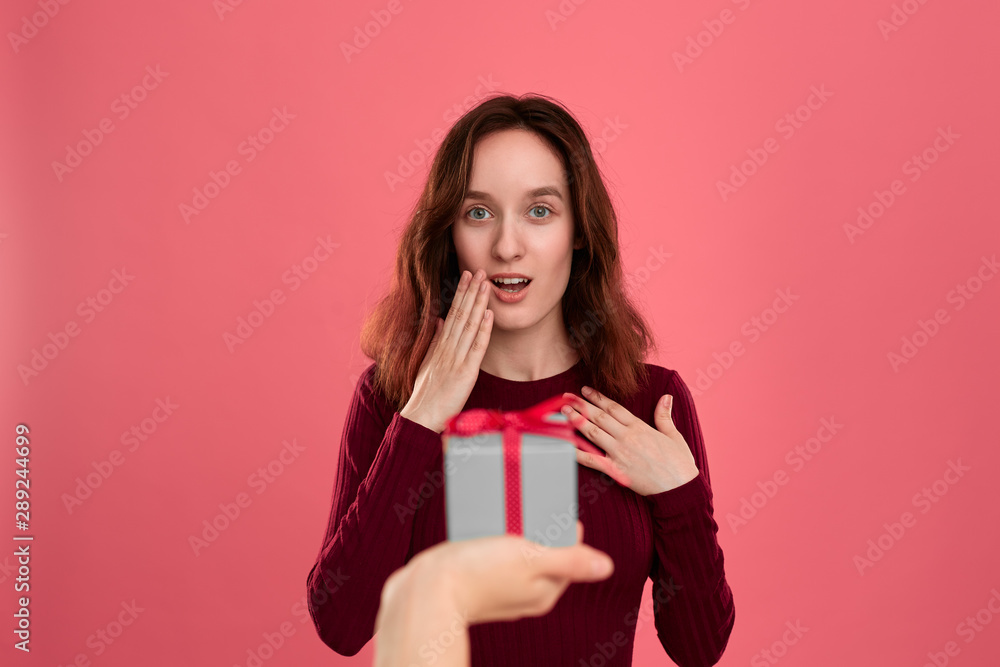 Happy pretty brunette girl gets a present box with a ribbon from another person standing isolated on a dark pink background and smiling at the camera. Celebrating special event. Happy pretty brunette girl gets a present box with a ribbon from another person standing isolated on a dark pink background and smiling at the camera. Celebrating special event.