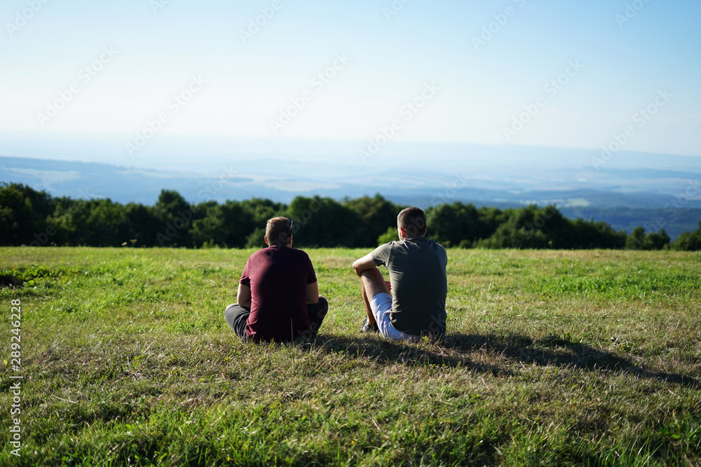 Gay couple or two friends sitting on the grassy hill and watching romantic sunset