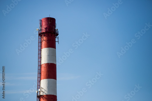 Red and white chemical plant pipe throws harmful poisonous smoke into clear blue sky with copy space. Concept of greenhouse effect and air pollution due to fuel combustion and chemical production.