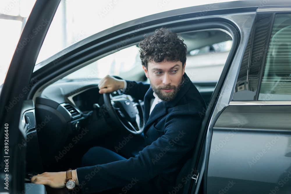 Portrait of young handsome man in his new silver gray car, relaxing, hand on steering wheel, looking out window, isolated on outdoors background with vehicle.