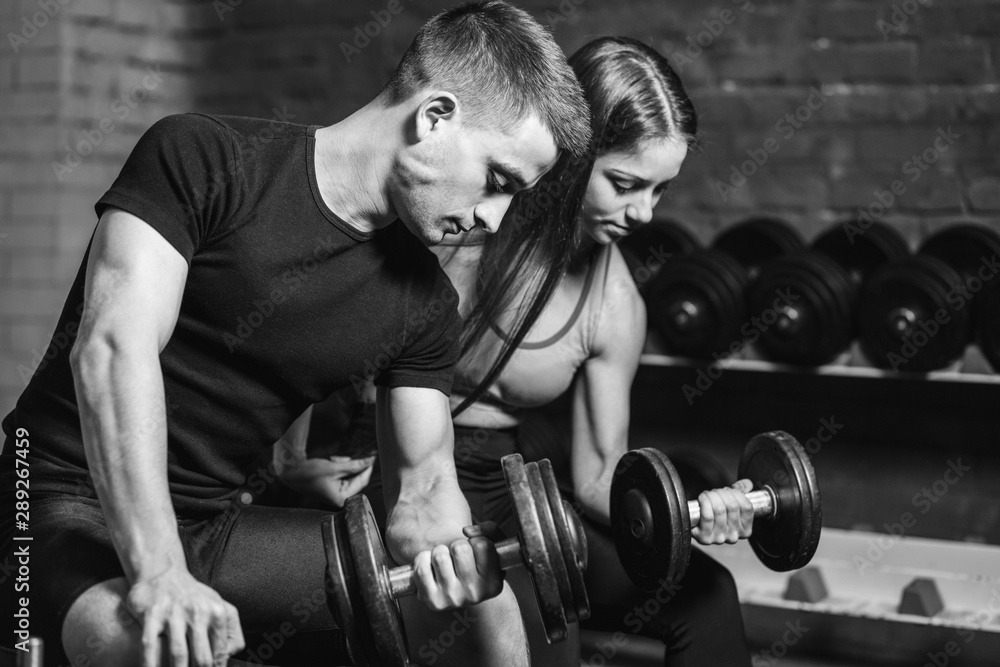 Fitness, sports, exercises and weightlifting. Concept - a young woman and a young man with dumbbells sweeping muscles in the gym. During this time they look at their muscles.