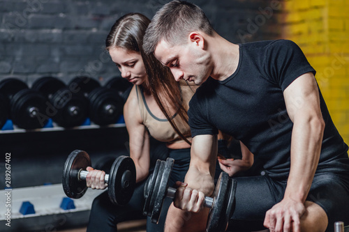 Fitness, sports, exercises and weightlifting. Concept - a young woman and a young man with dumbbells sweeping muscles in the gym. During this time they look at their muscles.