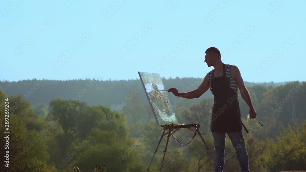 Young artist painting a landscape, day time. Landscape painter. Man painting a natural scenery with oil paints Young artist painting a landscape, day time. Landscape painter. Man painting a natural scenery with oil paints
