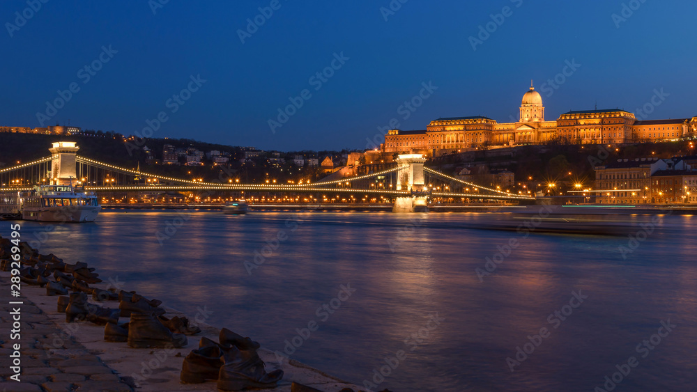 Panoramic view of Chain Bridge and Buda Castle on the west bank of the river Danube captured at the blue hour. (Shoes on the Danube Bank memorial in the foreground)