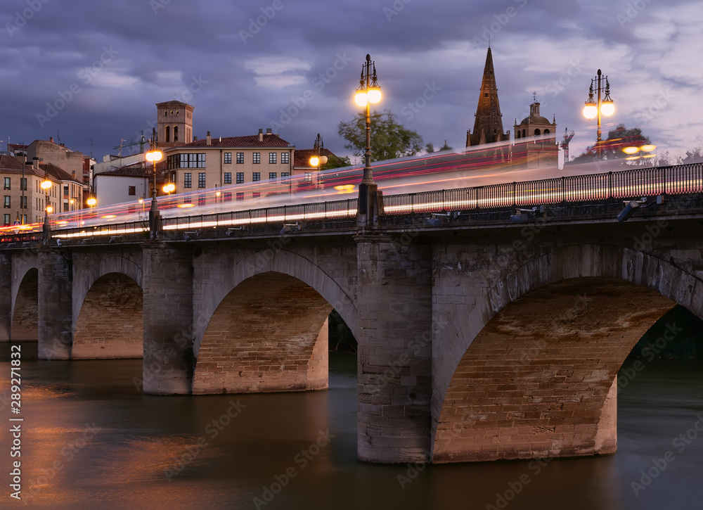 Fototapeta premium vistas del puente medieval, el río Ebro y la ciudad historica de Logroño en la Rioja,España, durante el anochecer, con estelas y reflejos de los vehiculos que transitaban el lugar