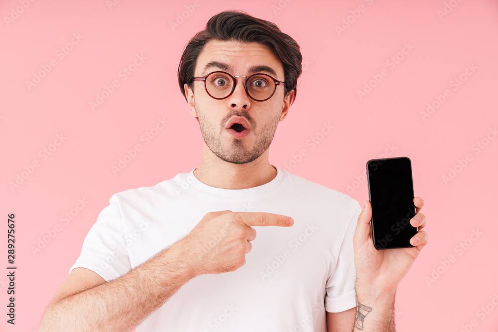 Image of unshaven surprised man wearing eyeglasses holding and pointing finger at cellphone Image of unshaven surprised man wearing eyeglasses holding and pointing finger at cellphone