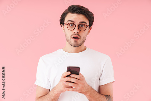 Image of unshaven shocked man wearing eyeglasses looking at camera and typing on cellphone