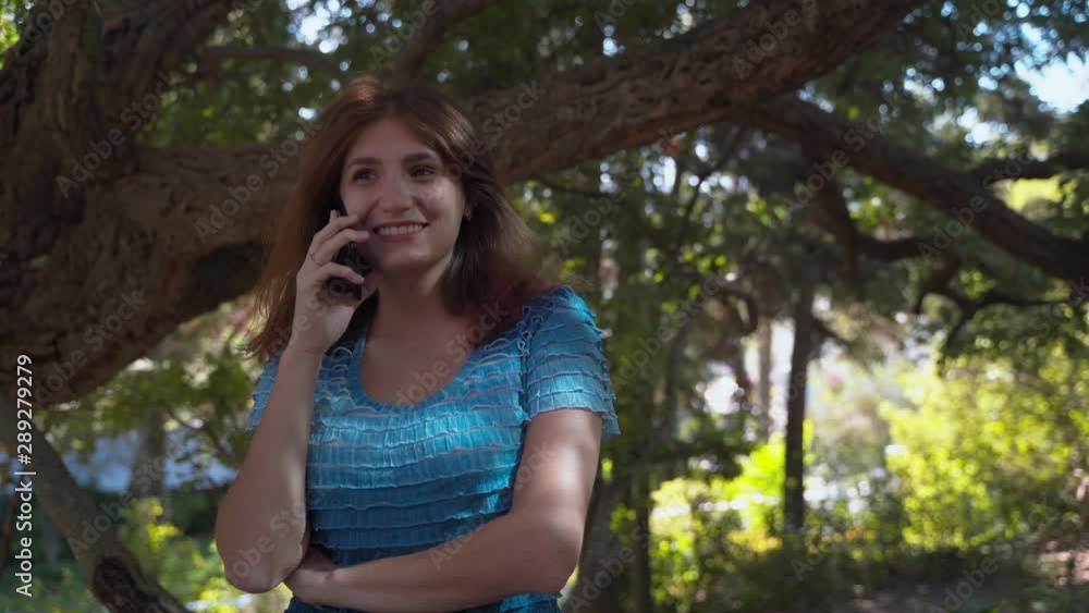 The girl in a blue dress happily speaks on the phone, smiles. Talking on the phone in the park. 4k The girl in a blue dress happily speaks on the phone, smiles. Talking on the phone in the park. 4k