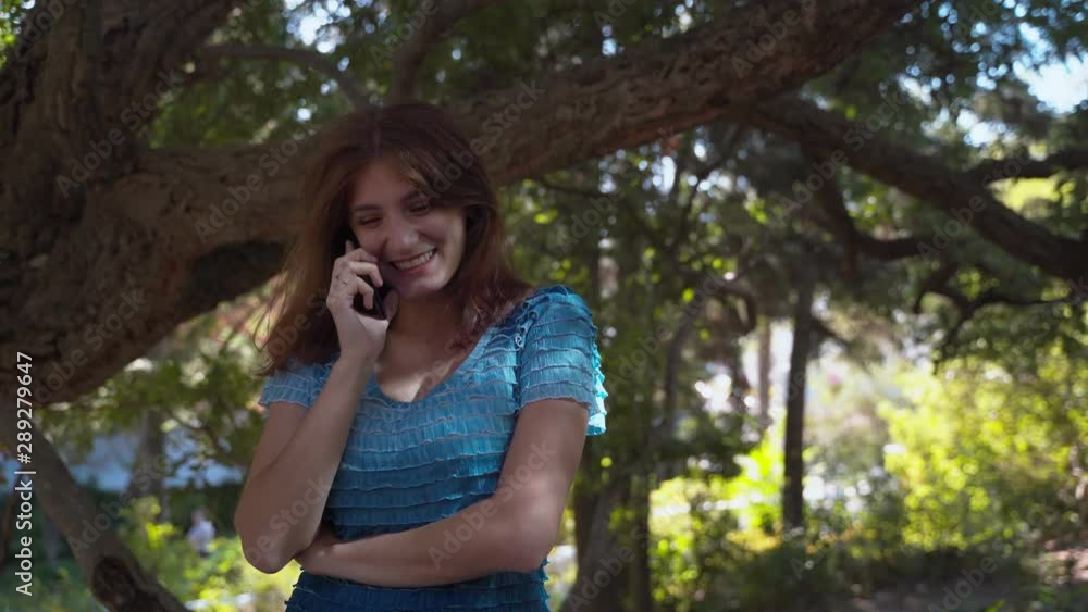 The girl in a blue dress happily speaks on the phone, smiles. Talking on the phone in the park. 4k The girl in a blue dress happily speaks on the phone, smiles. Talking on the phone in the park. 4k