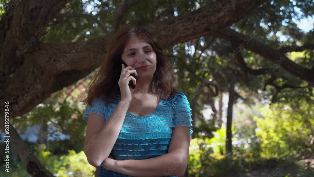 The girl in a blue dress happily speaks on the phone, smiles. Talking on the phone in the park. 4k The girl in a blue dress happily speaks on the phone, smiles. Talking on the phone in the park. 4k