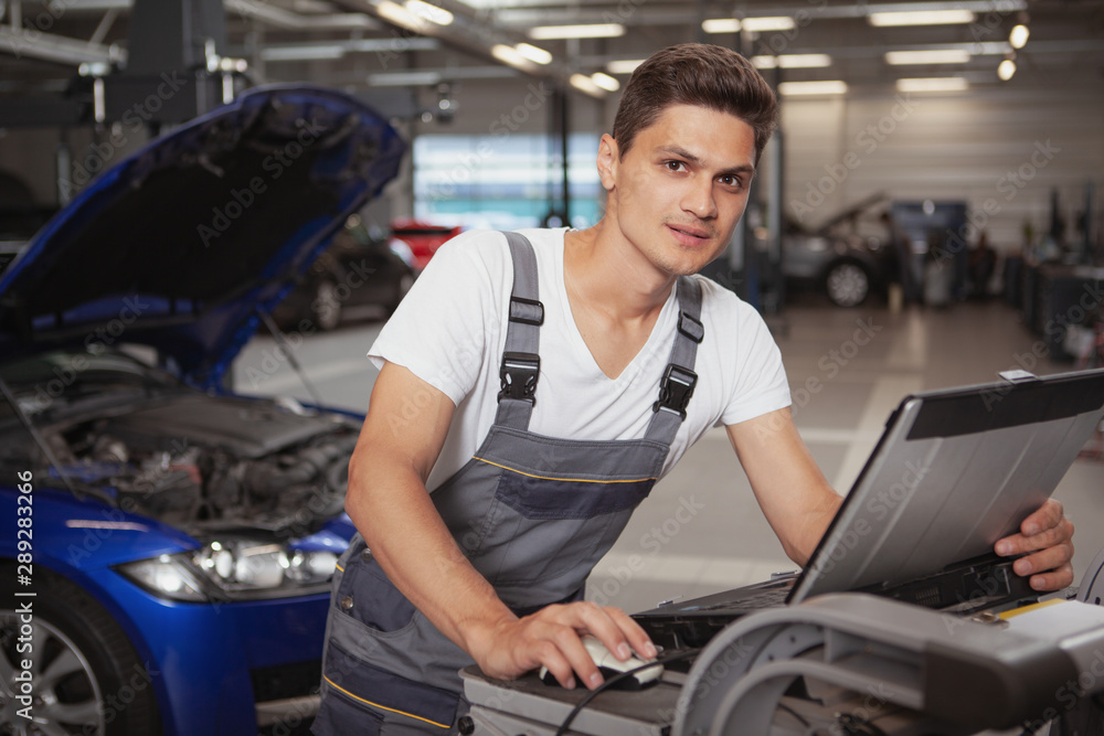 Young handsome car mechanic smiling to the camera, using laptop for computer diagnostics of a broken vehicle. Cheerful car technician working at the garage Young handsome car mechanic smiling to the camera, using laptop for computer diagnostics of a broken vehicle. Cheerful car technician working at the garage