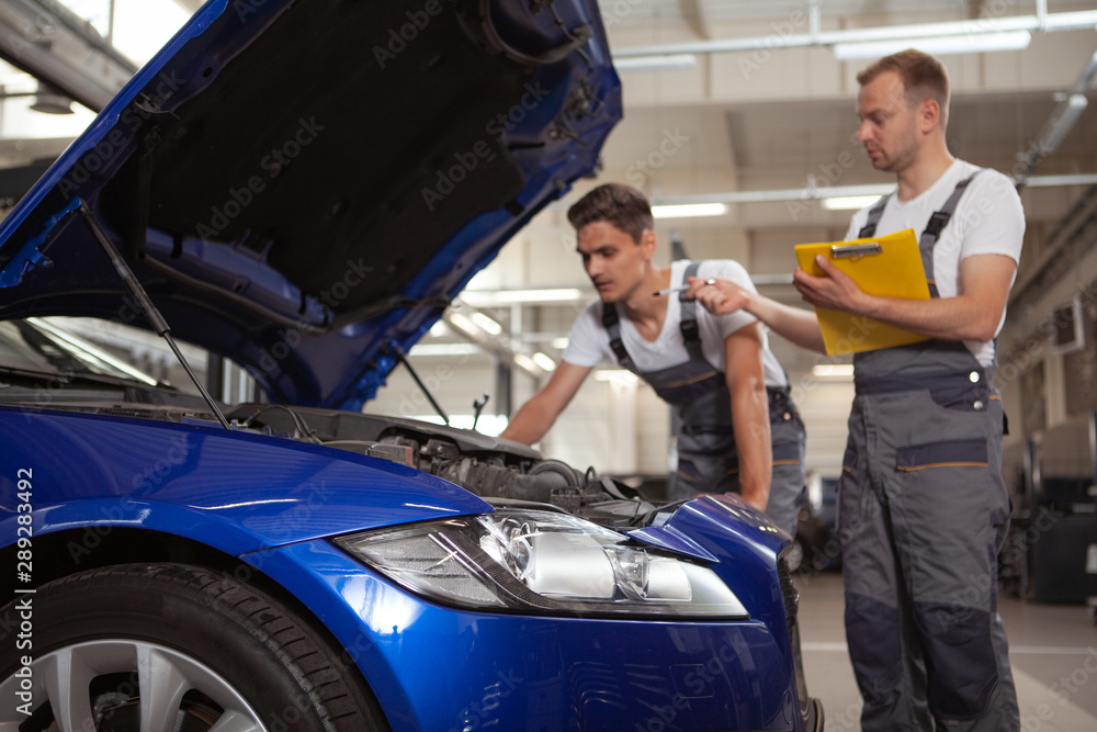 Selective focus on car lights, two mechanics examining engine of a car on background. Broken automobile at car service station Selective focus on car lights, two mechanics examining engine of a car on background. Broken automobile at car service station