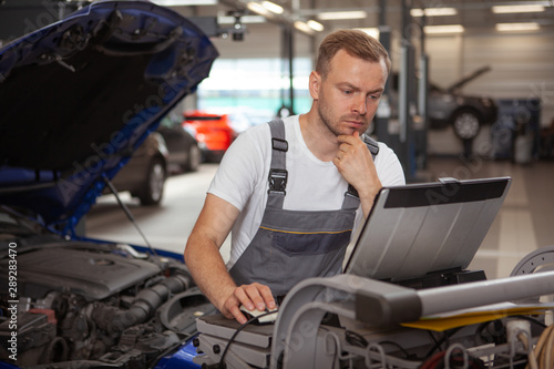Mature car mechanic looking focused, using laptop while repairing a car at the garage. Experienced auto technician using computer at his workshop