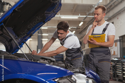 Two male car mechanics repairing broken automobile. Mature auto technician supervising his younger collegaue during automobile repair at the garage © Ihor