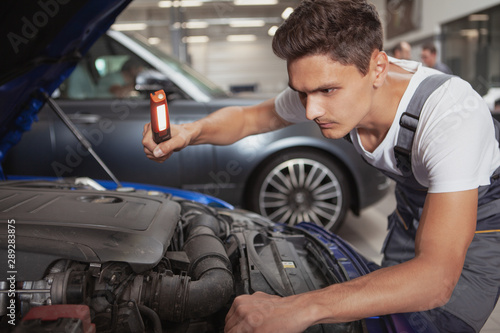 Young male car service worker looking under the hood of an automobile at the garage. Handsome auto mechanic examining engine of an auto at his workshop
