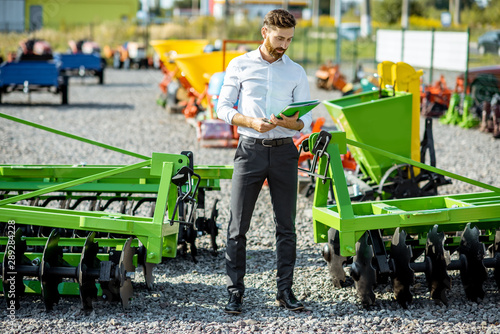 Portrait of a handsome salesman standing near the plow at the outdoor ground of the shop with new agricultural machinery © rh2010
