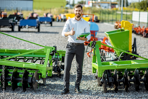Portrait of a handsome salesman standing near the plow at the outdoor ground of the shop with new agricultural machinery © rh2010