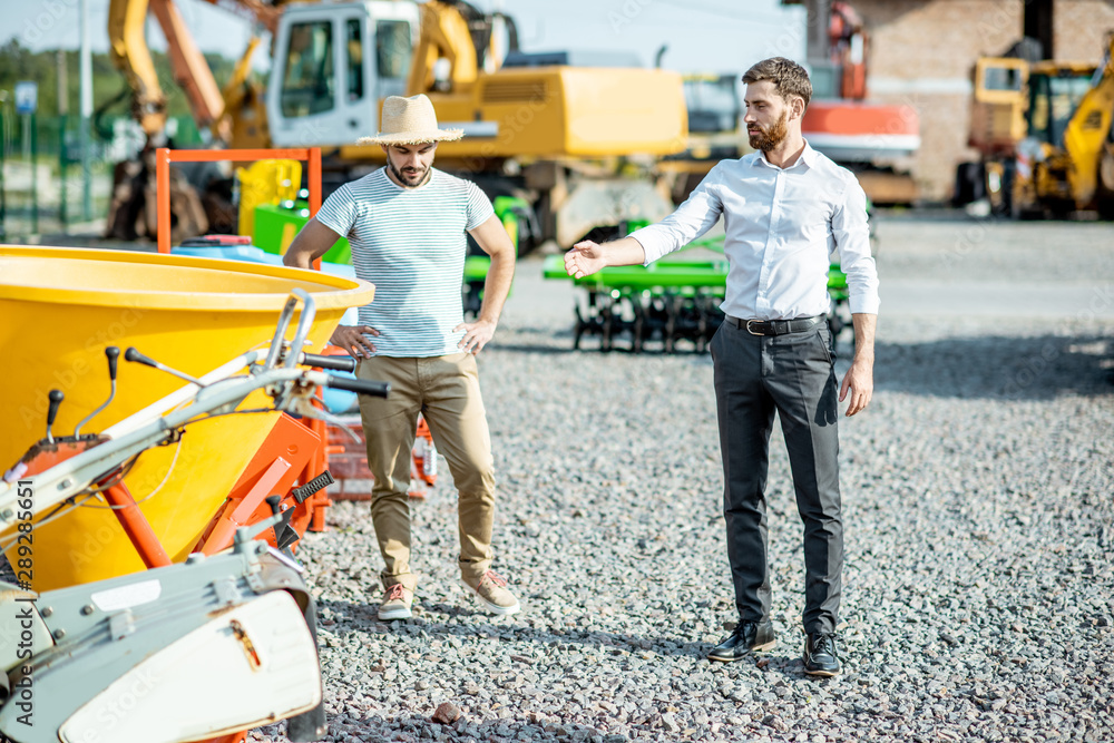 Young agronomist with salesman at the open ground of the shop with agricultural machinery, buying a new planter for farming Young agronomist with salesman at the open ground of the shop with agricultural machinery, buying a new planter for farming