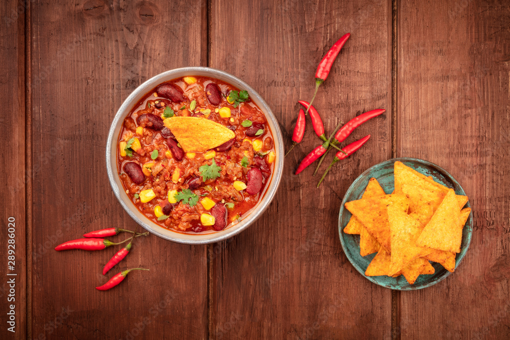 Chili con carne, a traditional Mexican dish with red beans, cilantro leaves, ground meat, and chili peppers, overhead shot with nachos on a dark rustic wooden background Chili con carne, a traditional Mexican dish with red beans, cilantro leaves, ground meat, and chili peppers, overhead shot with nachos on a dark rustic wooden background