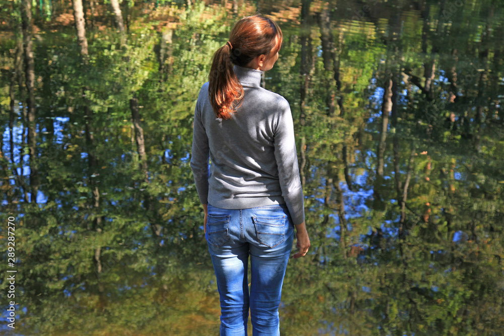 Young girl is standing near the pond and looking into the distance. Autumn morning landscape with woman and sun rays that reflected on the clear water. Pastime outdoors
