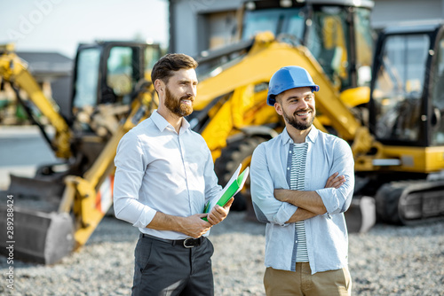 Builder choosing heavy machinery for construction with a sales consultant standing with some documents on the open ground of a shop with special vehicles photo