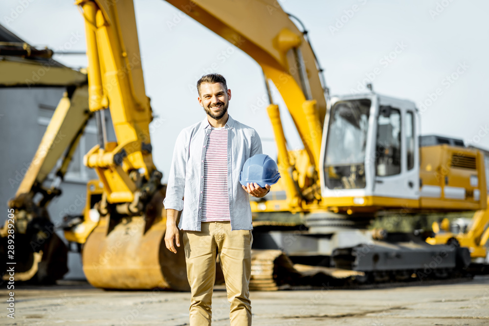 Portrait of a handsome builder standing on the open ground of the shop with heavy machinery for construction