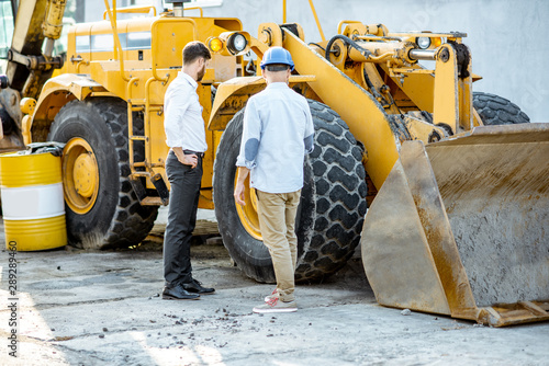 Builder choosing heavy machinery for construction, talking with a sales consultant on the open ground of a shop with special vehicles